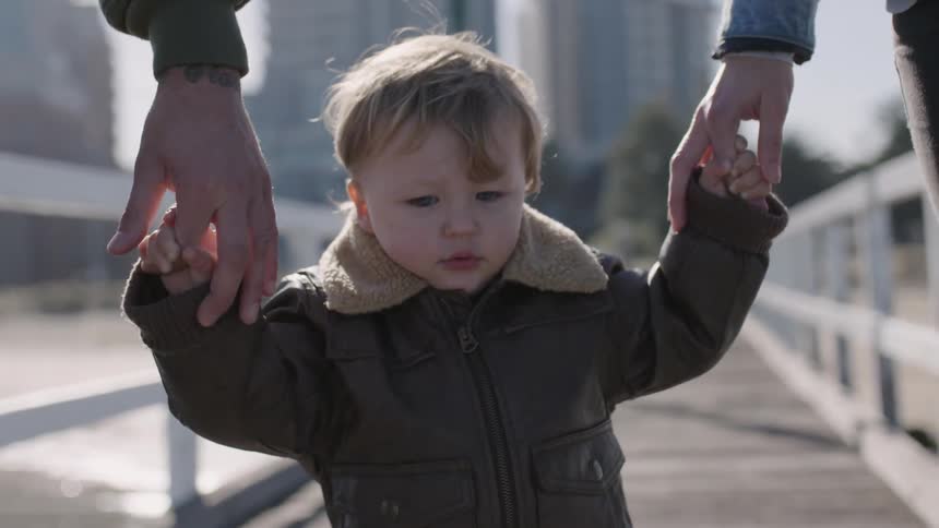 Two adults hold a toddler's hands walking on a bridge on a sunny day