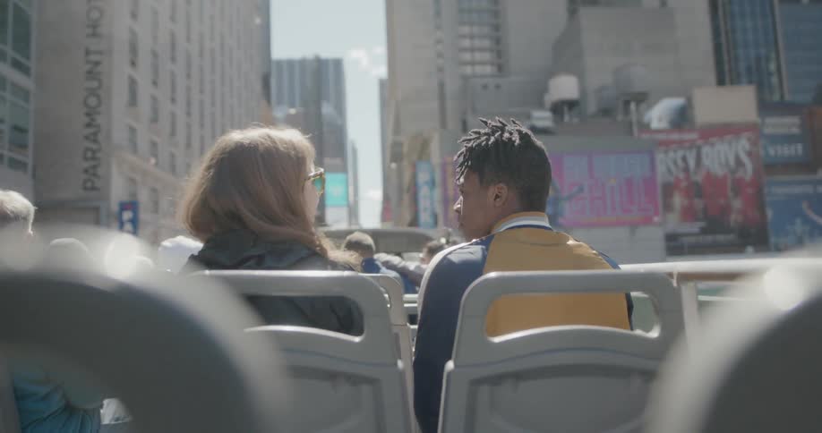 Man and woman sit on top of a tour bus in the city on a sunny day