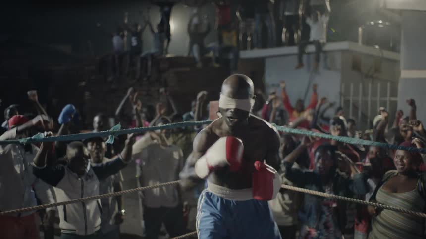 Blindfolded man stands in a boxing ring with an audience cheering behind him