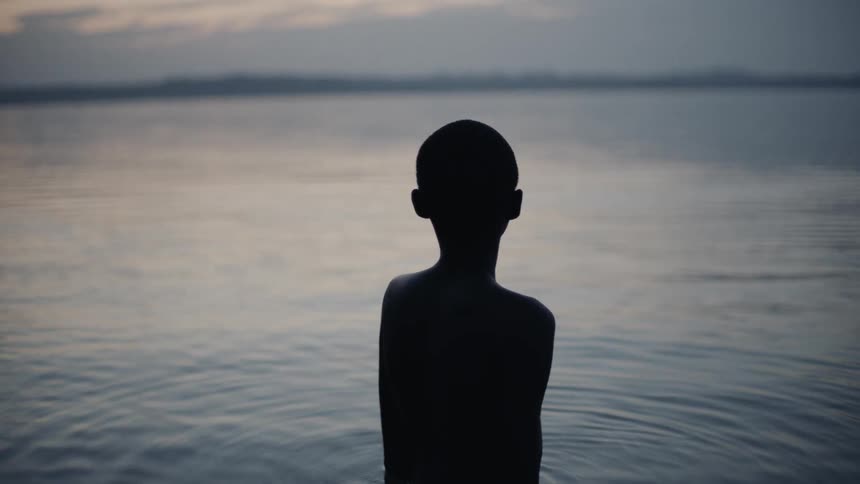 A boy is standing waist-deep in a lake at dawn