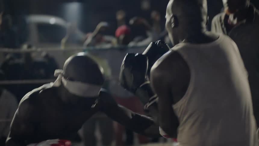 A blind-folded boxer is fighting another man in a ring outside at night