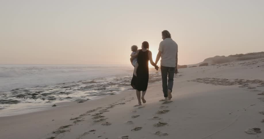 A couple hold hands as they walk on the beach at sunset with their baby