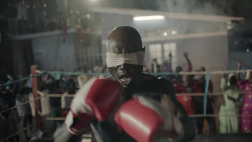 Blindfolded man punches in a boxing ring with an audience cheering behind him