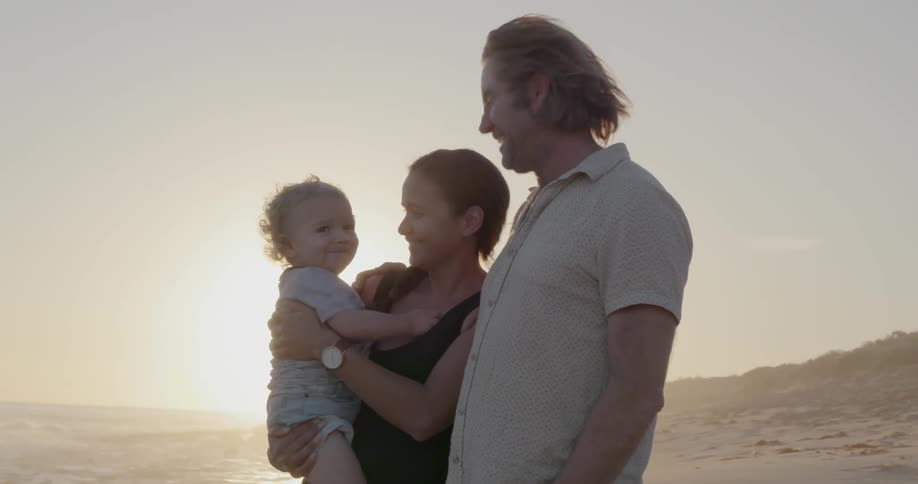 A couple are holding a baby outside on a beach at sunset