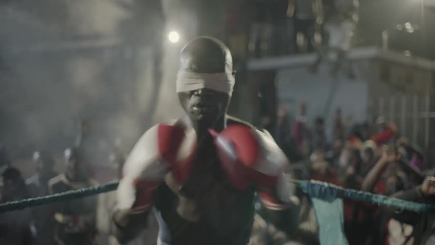 Blindfolded man stands in a boxing ring with an audience cheering behind him
