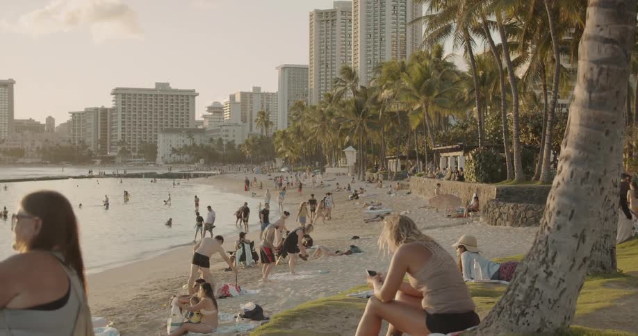 People walk and sit on a crowded beach near a city