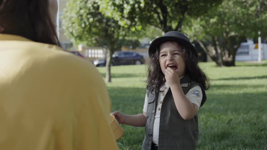 A boy is eating a bite of food as his mother watches outside on a sunny day