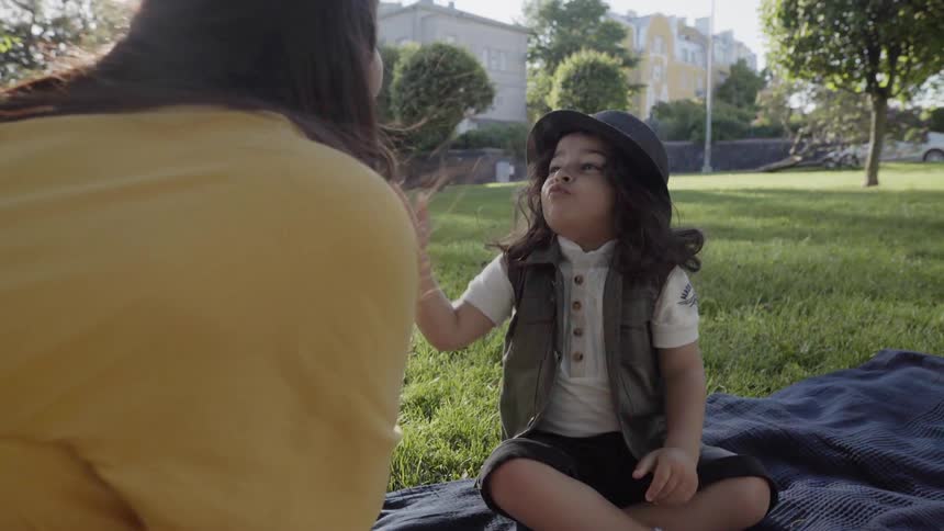 A child sitting on picnic blanket talking happily with mother in park.