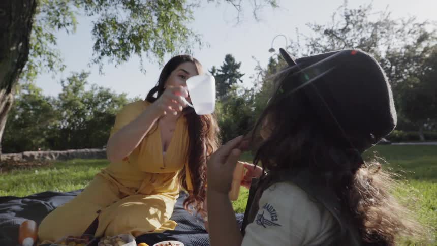 A mother drinking water from a cup while picnicking outdoors with child.