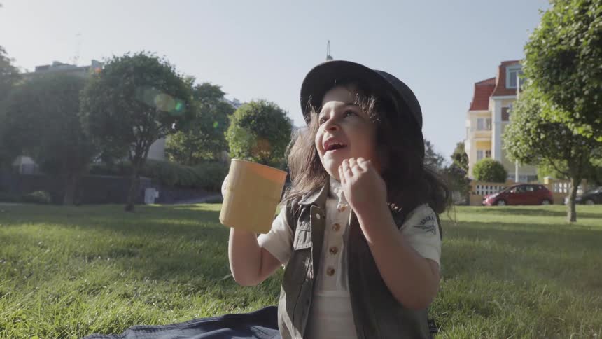 A cheerful child wearing a hat drinking from a cup and laughing outdoors.