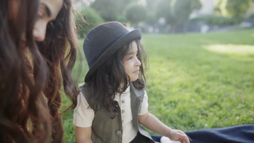 A young boy wearing a hat sitting with his mother during park picnic.