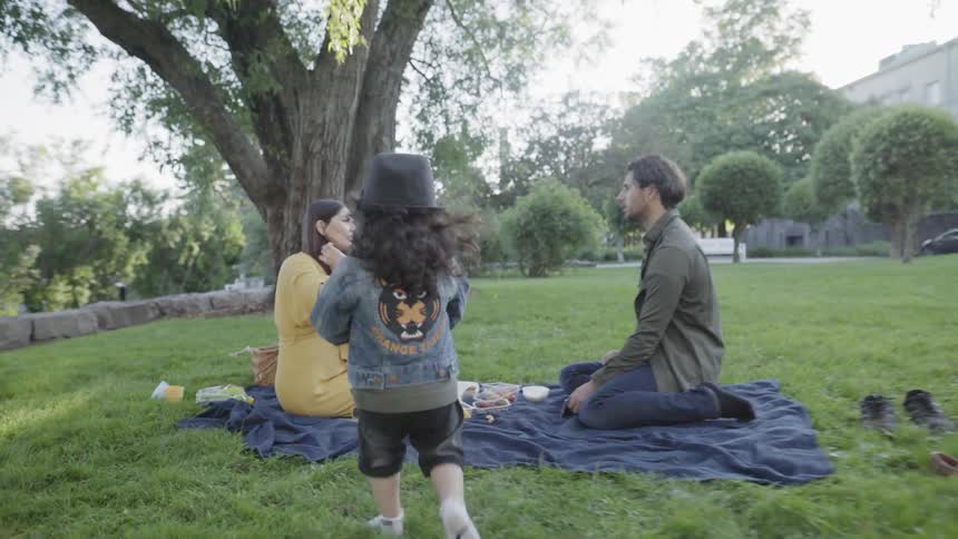 A young child wearing a hat walks towards parents sitting on picnic blanket.