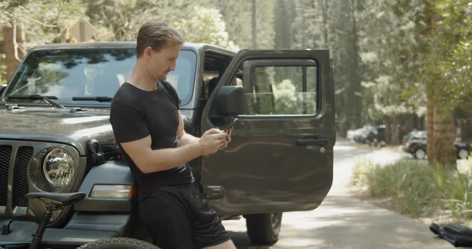 Man types on his phone next to a parked car