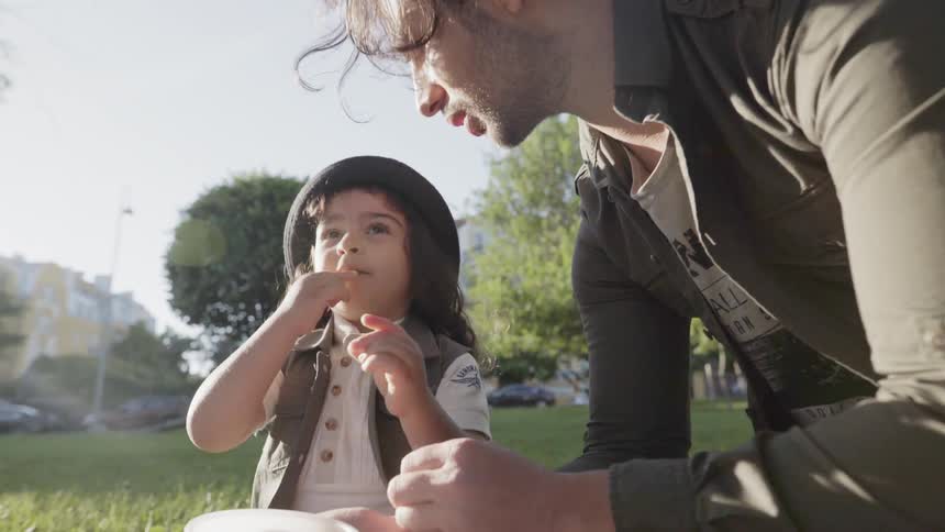 A father in an olive jacket feeding fruits to his young child at park.