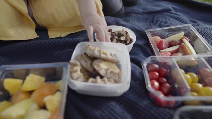 A young child reaches for fresh fruit containers during a family picnic.
