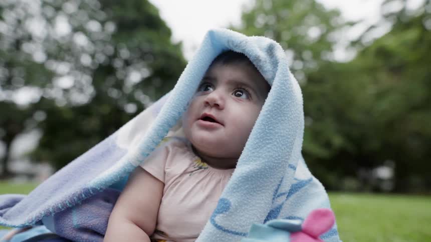 A baby sits wrapped in a blanket on a grassy lawn