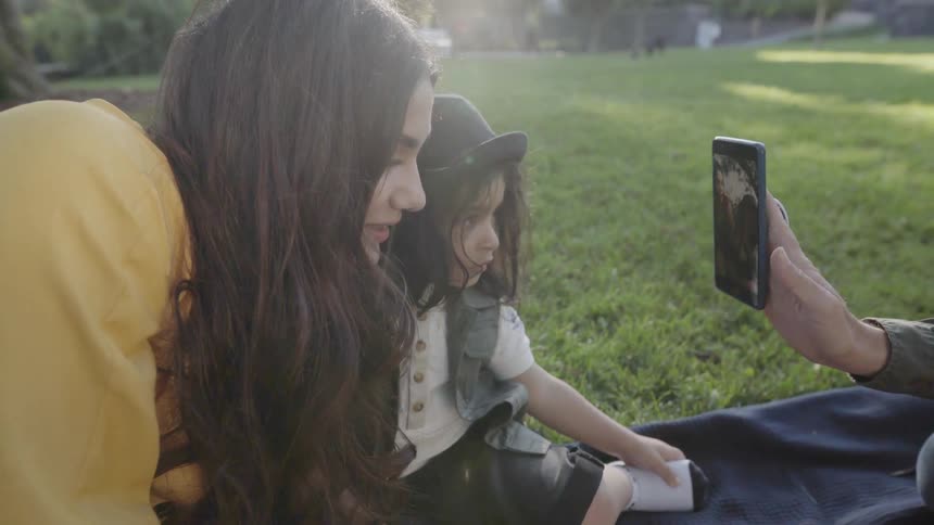 A man is showing a woman and boy photos on a cellphone as they sit in a park in daylight