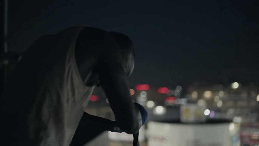 A man is leaning on a pole and then shadow boxing on a rooftop at night