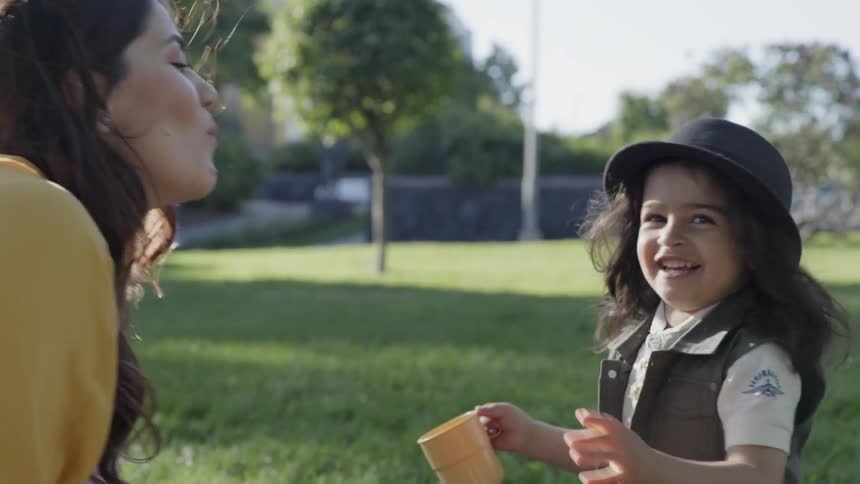 A young boy wearing a hat smiling brightly while talking to mother outdoors.