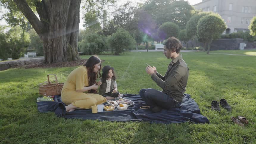 A young family sitting on a blanket taking pictures during park picnic.