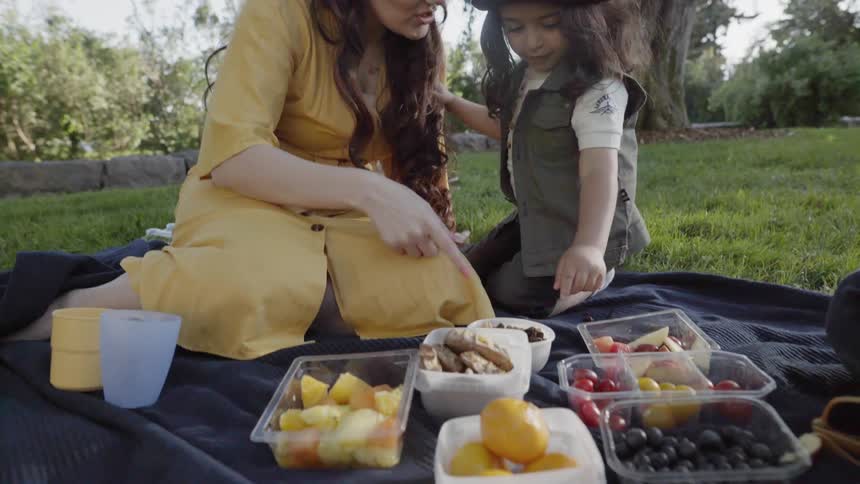 A mother points at fresh fruit snacks on blanket for her child.