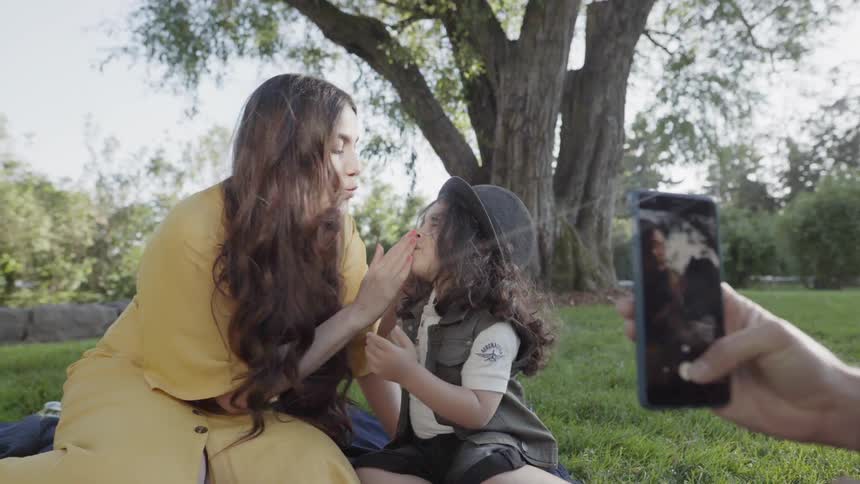 A mother playfully touching child face while father takes photo with smartphone.
