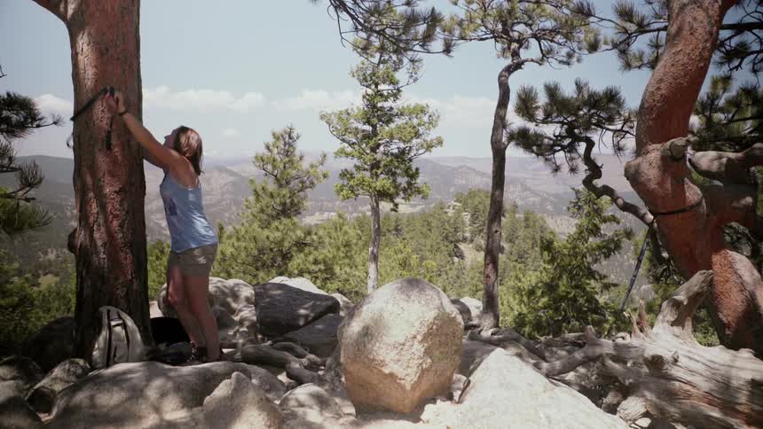 Woman ties a black rope around a tree on a hilltop by large rocks