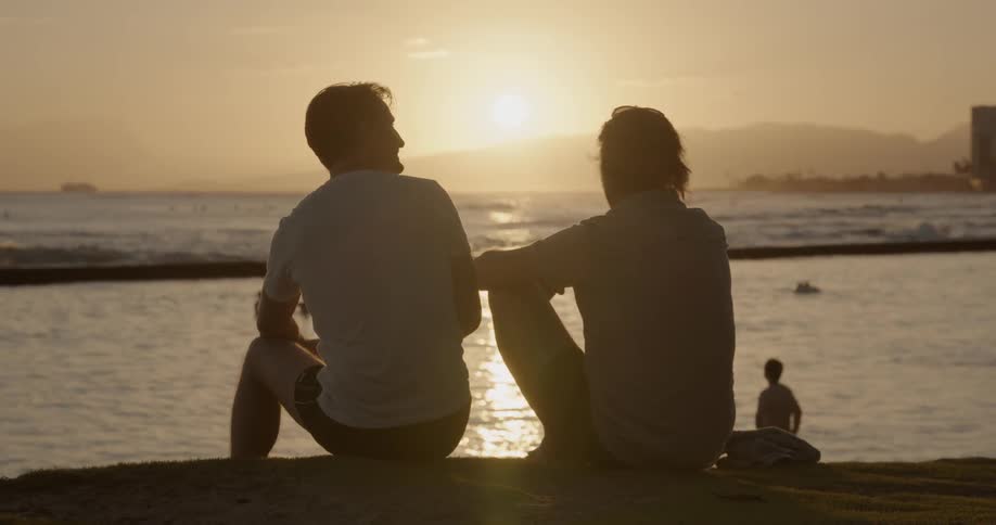 Two men sit on a beach at sunset