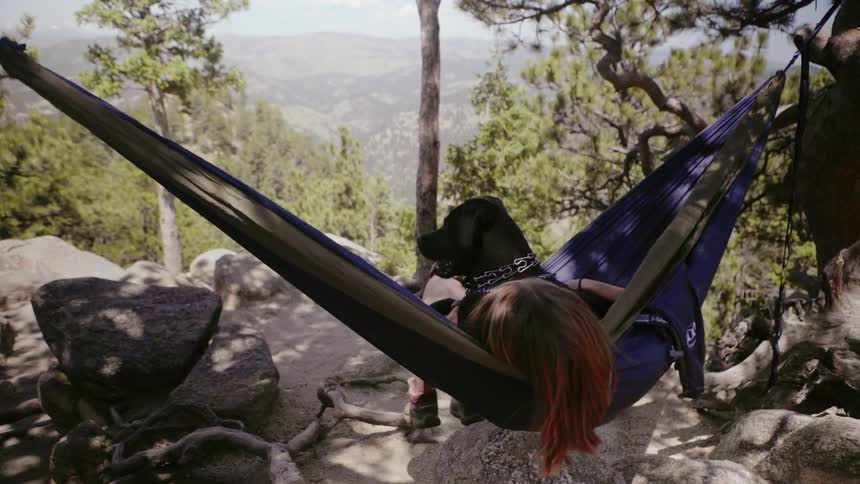 Woman and dog sit on a hammock on a rocky hilltop