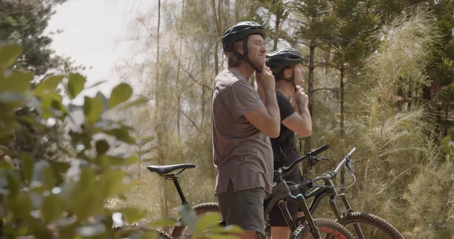 Two men on bikes in the woods put their helmets on and ride away