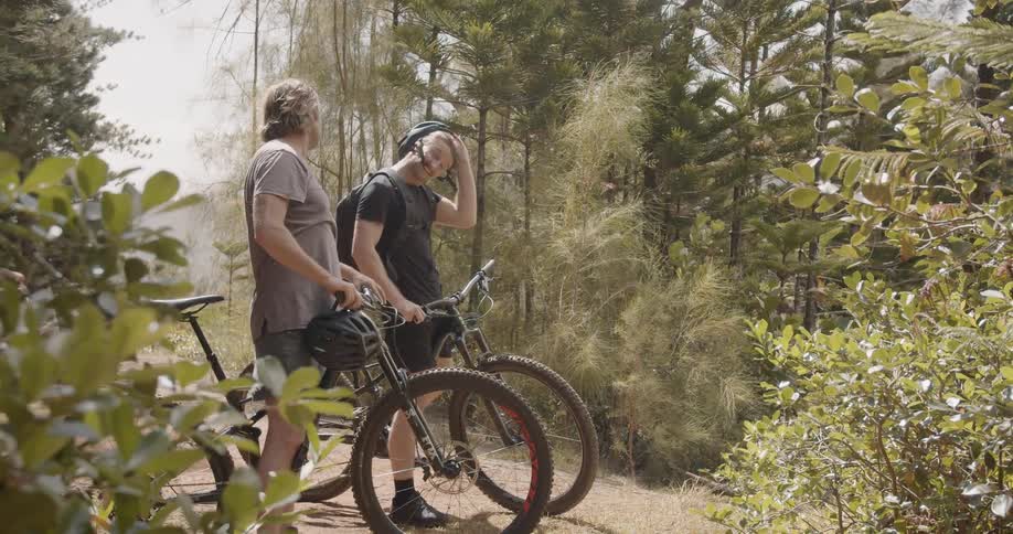 Two men ride bikes in the woods and stop and take off their helmets