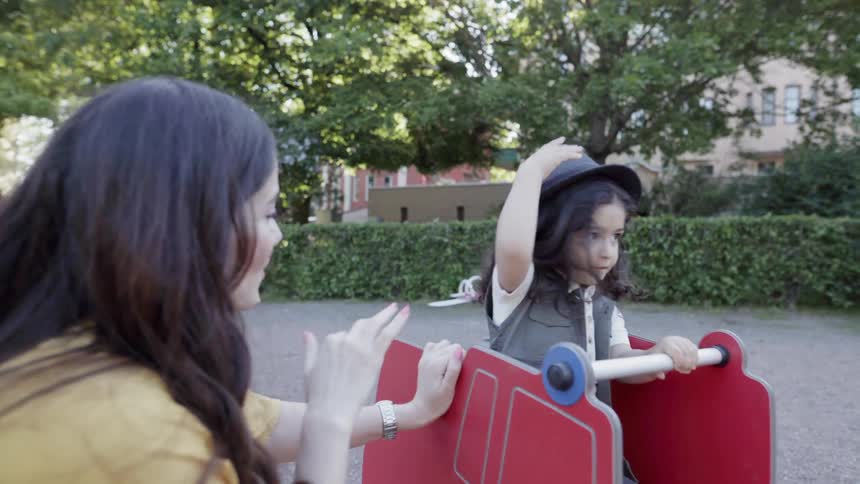 Child rides on park equipment and a woman talks to him