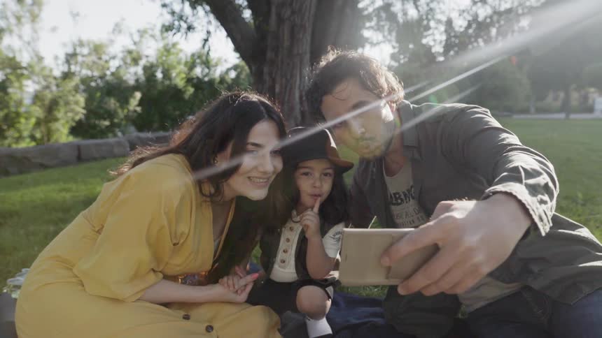 A young family posing together taking a selfie photo on smartphone in park.