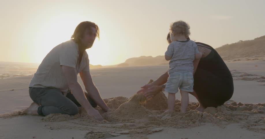 A young family builds a sandcastle on a beach at sunset