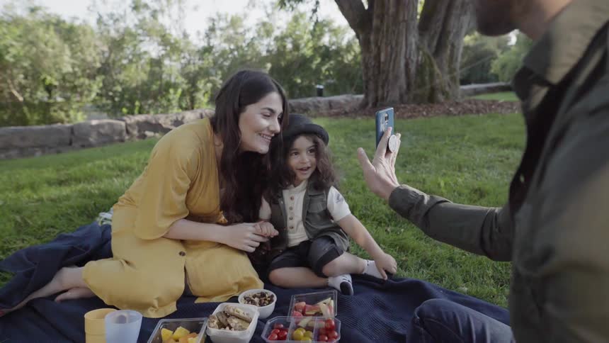 A man is showing a woman and boy photos on a cellphone as they sit in a park in daylight