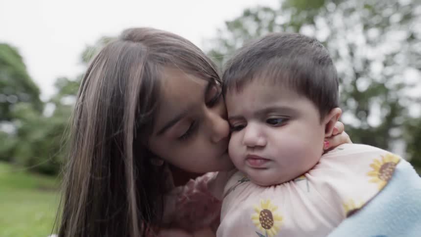 A girl kisses a baby while sitting on a grassy lawn