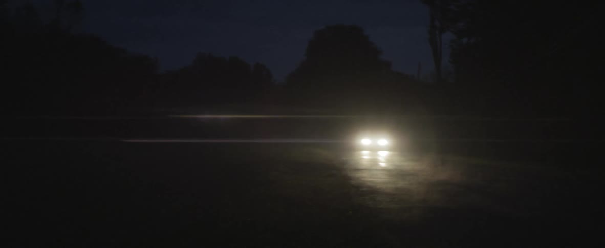 A car with its headlights on travels down a dark road