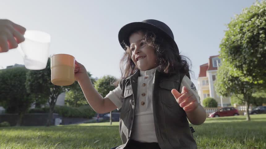 A happy young child clinking plastic cups with a parent during park picnic.
