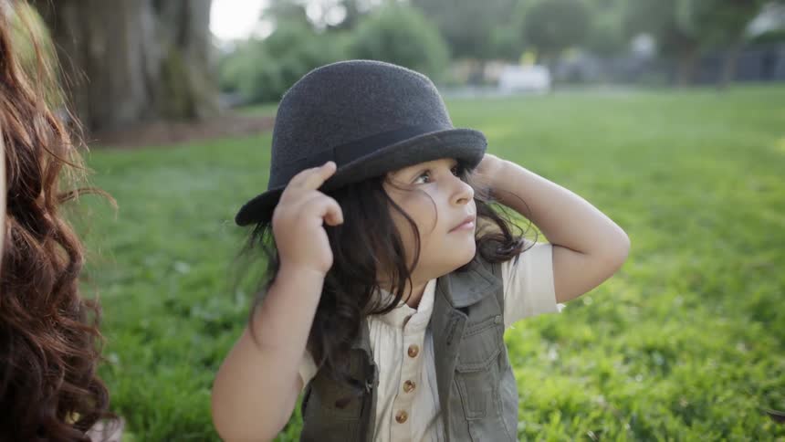 A little boy is adjusting the hat on his head as he picnics with her mother in daylight