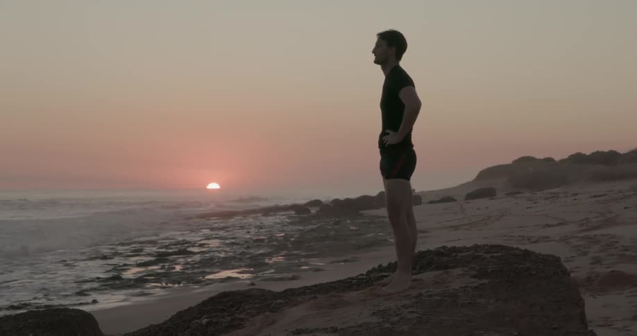 A man is standing on a beach at sunset looking out at the waves
