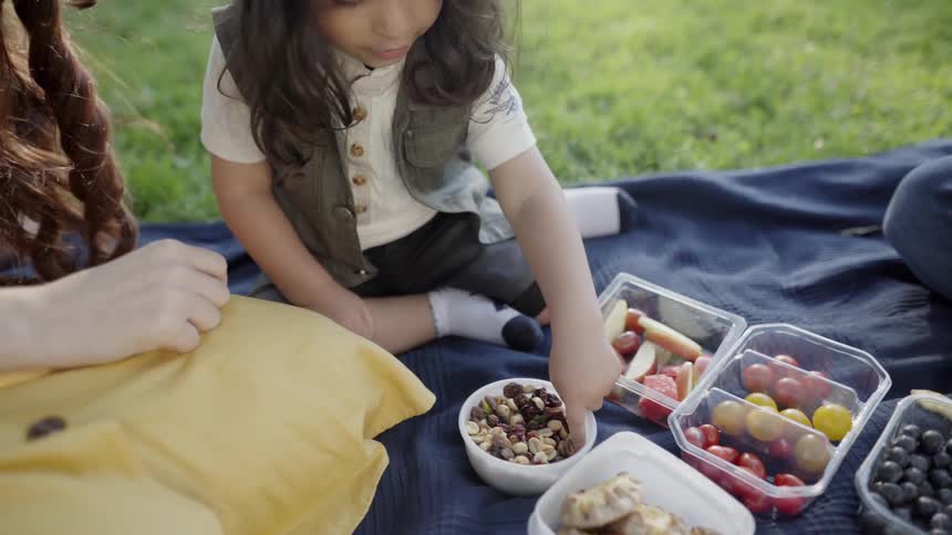 A young child reaches for healthy snacks on blanket during park picnic.