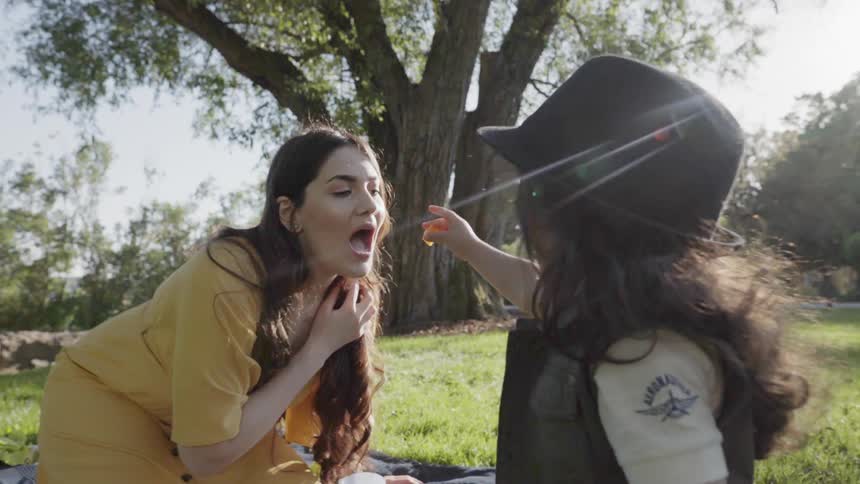 A playful child feeding a snack to mother during a family park picnic.