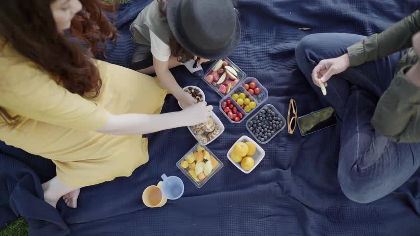 A family sitting together on a dark blanket sharing fruit picnic outdoors.