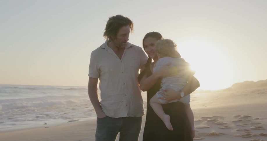 A man and woman are standing holding their child on a beach at sunset