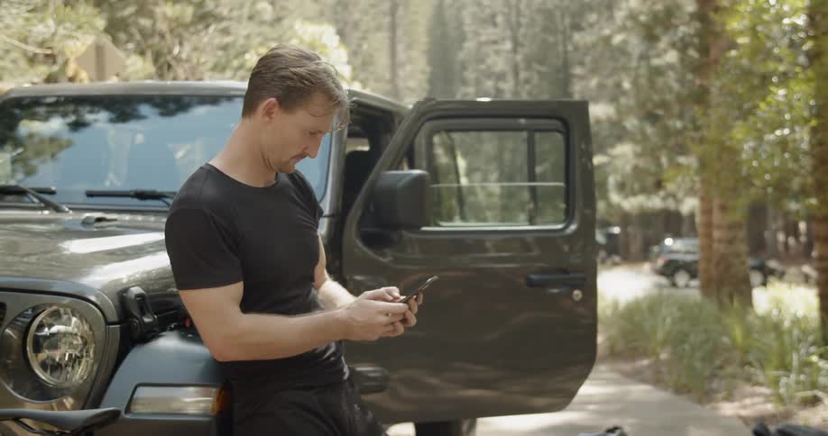 A man types on his phone while leaned against a car