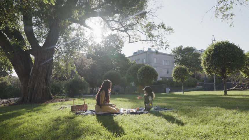 A young child sitting across from mother on a blanket in park.