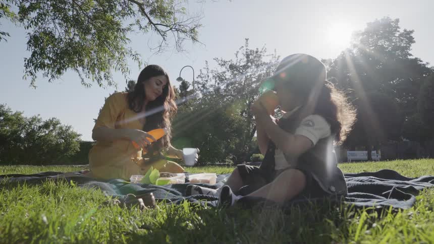 A young child drinking water while mother prepares snacks on picnic blanket.