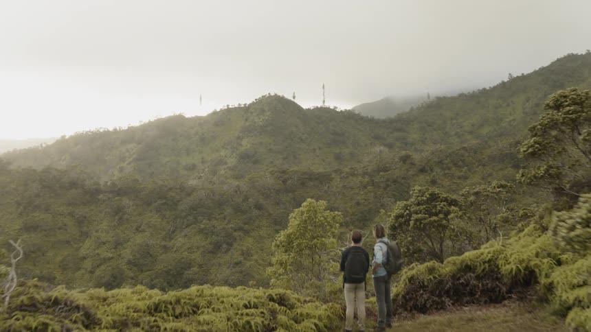 Two people stand on a grassy hilltop looking out at the hills