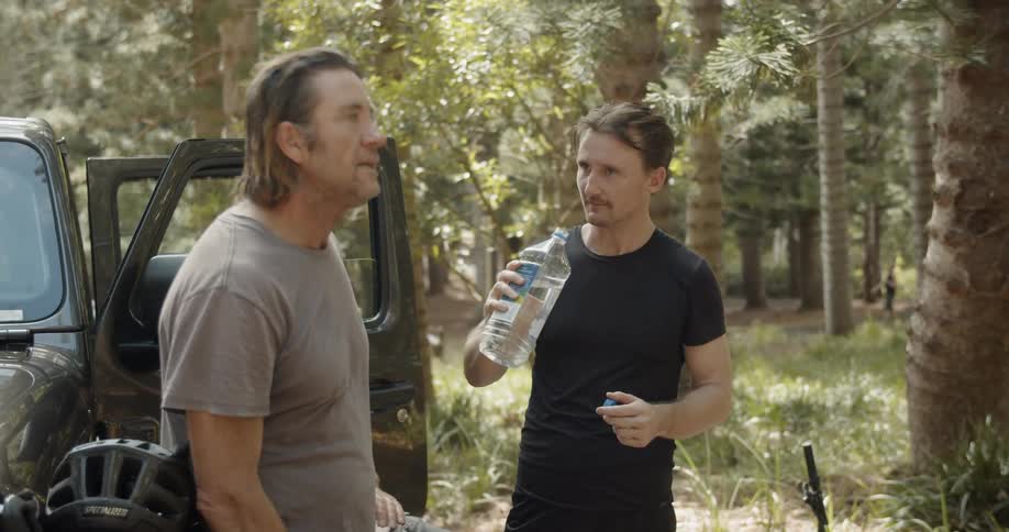 Two men talk next to a parked car in a forest
