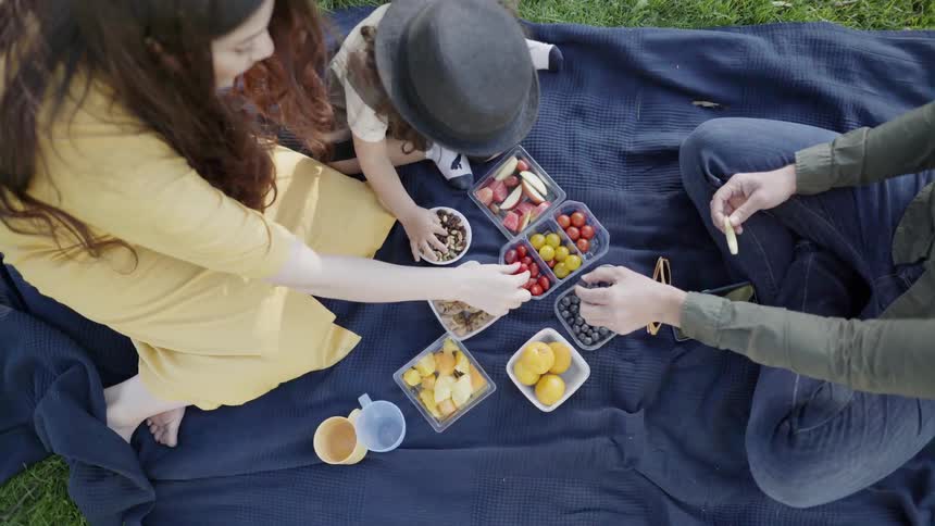A family sitting on a blue blanket sharing fruit and snacks.